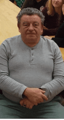 a man in a grey shirt is sitting with his hands folded in front of a group of people