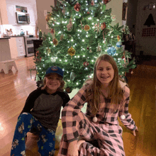 a boy wearing a nasa hat sits next to a girl next to a christmas tree