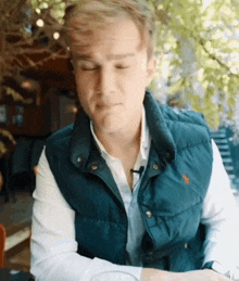 a young man wearing a blue vest and a white shirt is sitting at a table with his eyes closed .