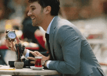 a man in a suit is sitting at a table with a cup of tea and a clock in his hand