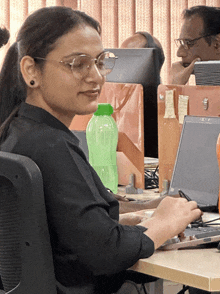 a woman wearing glasses sits at a desk with a laptop and a green bottle