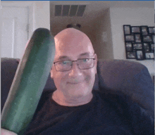 a bald man with glasses is holding a large cucumber in front of his face