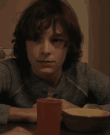 a young boy sits at a table with a bowl of food and a red cup