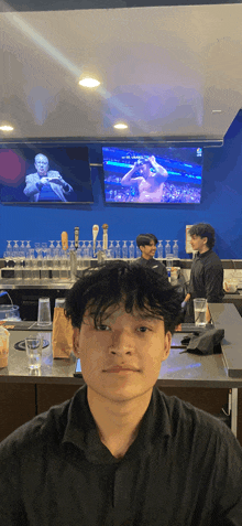 a man in a black shirt stands in front of a bar with a boxing match on the screen behind him