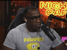 a man wearing a pawpaw shirt is sitting in front of a neon sign that says night cafe