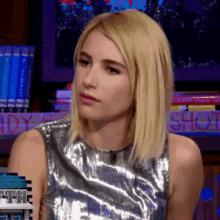 a woman in a silver top is sitting in front of a bookshelf with the word shot written on it