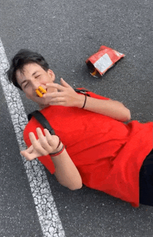 a young man in a red shirt is laying on the ground with a bag of chips on the ground