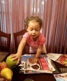 a little girl is playing with an apple on a table with a newspaper that says chocolate cake on it