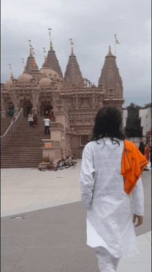 a man in a white shirt and orange scarf walks in front of a large temple