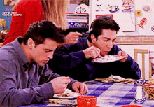 two men are sitting at a table with plates of food in front of a refrigerator that says " international gourmet "