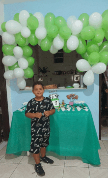 a boy stands in front of a table decorated with green and white balloons and a cake with the number 7 on it