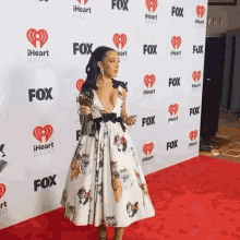 a woman in a white dress stands on a red carpet in front of a wall that says iheart radio