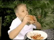 a young boy is eating a hamburger while sitting at a table with a plate of food in front of him