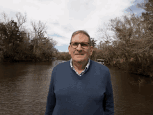 a man wearing glasses and a blue sweater stands in front of a body of water