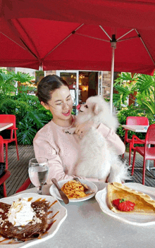 a woman petting a white dog while sitting at a table with food