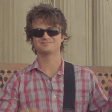 a man wearing sunglasses and a plaid shirt makes a heart shape with his hands