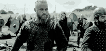 a man with a beard is standing in front of a group of vikings in a field .