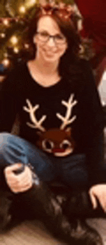 a woman wearing a black reindeer sweater is sitting on the floor in front of a christmas tree .