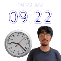 a man with glasses stands next to a clock that says 9:22 am