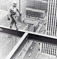 a black and white photo of a man standing on top of a metal beam overlooking a city .