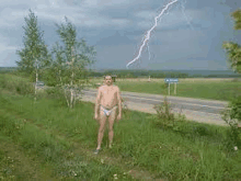 a shirtless man is standing in the grass with a lightning strike in the background .