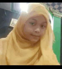 a young girl wearing a yellow hijab looks at the camera in front of a green wall