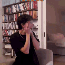 a man in a black shirt is sitting in front of a bookshelf and covering his face with his hands .