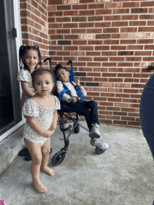 two little girls and a boy in a stroller are standing in front of a brick wall