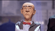 a bald puppet wearing a bow tie and a suit is standing with his arms crossed .