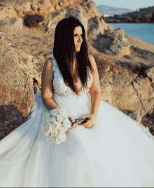 a woman in a wedding dress sits on a rock holding a bouquet of flowers