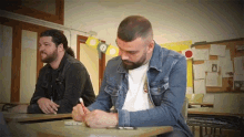 a man in a denim jacket sits at a desk in front of a bulletin board with the number 1 on it