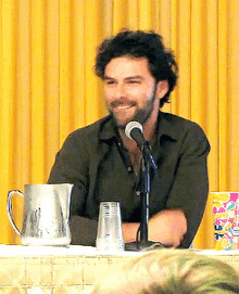 a man with a beard is sitting at a table with a pitcher and a glass in front of a microphone