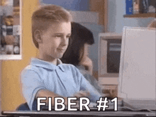 a young boy is sitting at a desk in front of a computer with the words fiber # 1 written on the screen .