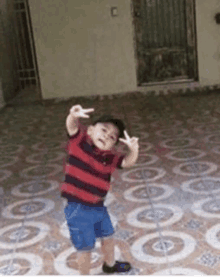 a little boy in a red and black striped shirt is standing on a tiled floor giving the peace sign