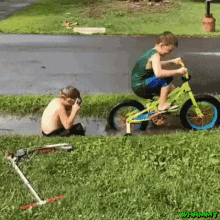 a boy is riding a bike in a puddle while another boy sits in the grass taking a picture .