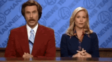 a man and a woman are sitting at a news desk in front of a channel 4 news wall .