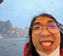 a woman wearing glasses and a face mask smiles in front of a snowy street