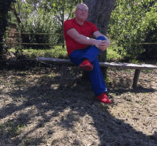 a man in a red shirt and blue pants sits on a wooden bench