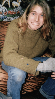 a man with long hair wearing a green sweater and jeans smiles for the camera