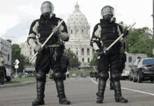 two riot police officers are standing next to each other on a city street holding batons .