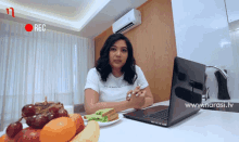 a woman sits at a table with a laptop and a plate of fruit with a red rec sign above her head