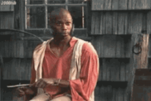 a man in a red shirt and white vest is sitting in front of a wooden building holding a gun .