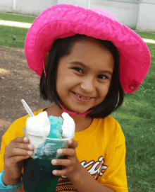 a young girl wearing a pink hat and a yellow shirt holds a cup of ice cream