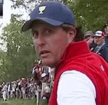 a man wearing a blue hat and a red vest is standing in front of a crowd of people on a golf course .