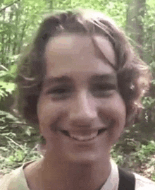 a young man with long hair is smiling for the camera while standing in the woods .