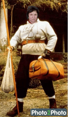 a woman wearing a 1972 olympics sweatshirt holds a bag and a pole