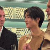 a man in a tan sweater holds a glass of champagne in front of a sign that says taehu
