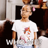 a little girl wearing glasses and a white shirt is sitting on a couch and making a funny face .