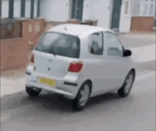 a small white car is driving down a street next to a house .