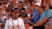 a young boy wearing a hat with the word angels on it is sitting in a crowd of people at a baseball game .